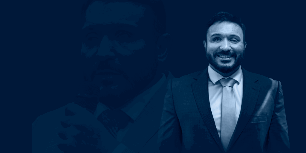 Nesta foto está Marcio Germano de terno e gravata, sorrindo, com barba curta e cabelo escuro. O fundo da imagem é totalmente azul escuro, e a imagem tem um filtro azul sobreposto, dando um tom monocromático. À esquerda, aparece parcialmente o contorno ampliado de sua orelha e rosto, como uma sobreposição da mesma imagem.